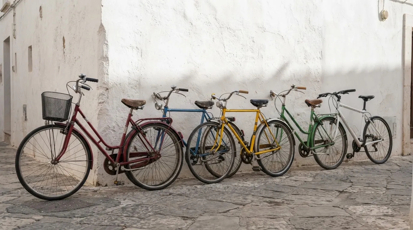 Fila de cinco bicicletas de cores diferentes encostadas a um muro caiado, vistas de lado, luz da manhã.
